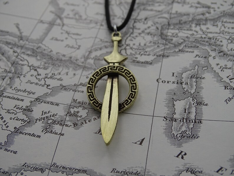 Może przedstawiać: Złoty naszyjnik z wisiorem w kształcie miecza, ozdobiony dekoracyjnym kołem. Koło ma wz&oacute;r greckiego klucza. Naszyjnik jest na czarnym sznurku i jest wyświetlany na starej mapie.