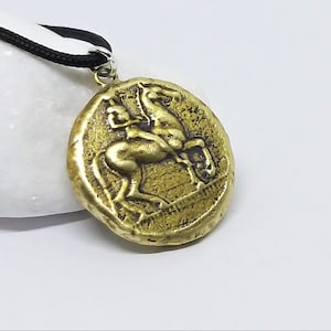 Puede incluir: Un collar con un colgante en forma de moneda griega antigua. La moneda es de color dorado y representa a un jinete a caballo, con detalles intrincados y una superficie texturizada.