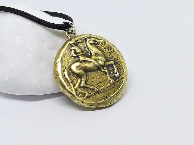 Puede incluir: Un collar con un colgante en forma de moneda griega antigua. La moneda es de color dorado y representa a un jinete a caballo, con detalles intrincados y una superficie texturizada.