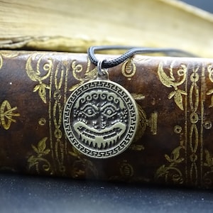 May include: A silver pendant necklace with a circular design featuring a stylized face with intricate details. The pendant is on a black cord and is resting on a brown leather-bound book with gold accents.