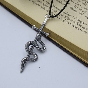 Op de afbeelding: Een zilveren ketting met een hanger met een slang die om een zwaard is gewikkeld. De hanger zit aan een zwart koord en ligt op een boek.