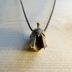May include: A gold-toned pendant necklace featuring a detailed Spartan helmet design. The pendant is attached to a black cord.