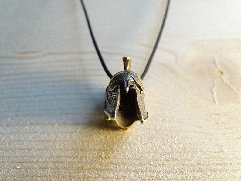 May include: A gold-toned pendant necklace featuring a detailed Spartan helmet design. The pendant is attached to a black cord.