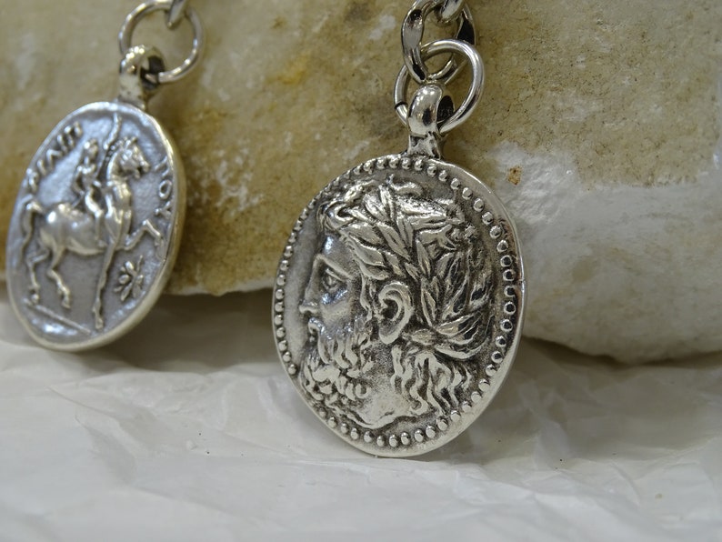 May include: Two silver-toned pendants with chain links. One pendant features a profile of a man with a beard and a laurel wreath. The other pendant features a horse and rider.