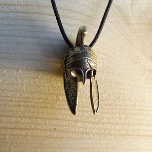 May include: A bronze pendant necklace featuring a detailed depiction of a Roman soldier's helmet. The helmet has a distinctive crest and a plume, and the pendant is suspended from a black cord.