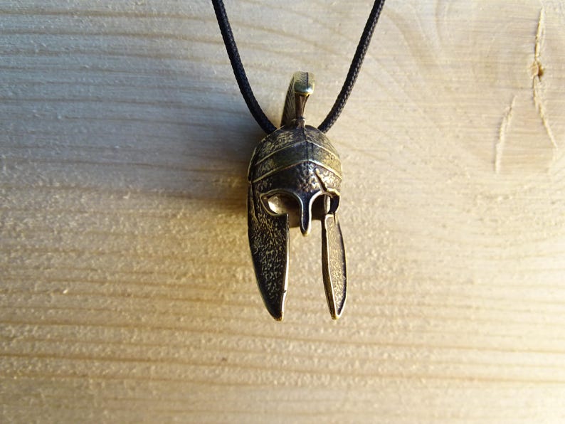 May include: A bronze pendant necklace featuring a detailed depiction of a Roman soldier's helmet. The helmet has a distinctive crest and a plume, and the pendant is suspended from a black cord.