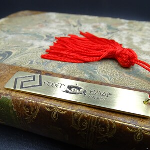 May include: A brass bookmark featuring a Greek design and the text "FREE'T'S HM&Delta;P OMHP&Theta;E". The bookmark is attached to a red tassel with a black bead.