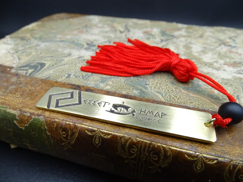 May include: A brass bookmark featuring a Greek design and the text "FREE'T'S HM&Delta;P OMHP&Theta;E". The bookmark is attached to a red tassel with a black bead.