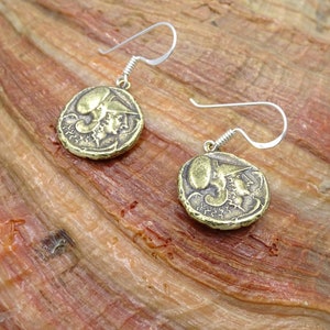 May include: A pair of gold-toned earrings with a coin-shaped design featuring a profile of a person's face. The earrings have silver hooks.