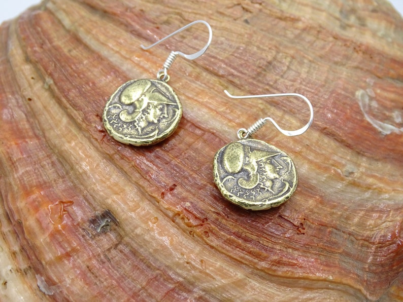 May include: A pair of gold-toned earrings with a coin-shaped design featuring a profile of a person's face. The earrings have silver hooks.