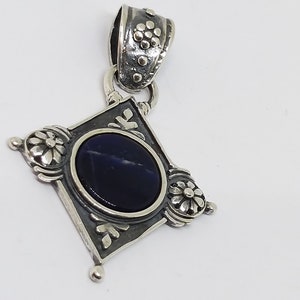 May include: A silver pendant with a dark blue gemstone set in a square frame with floral details. The pendant has a silver bail with a floral design.