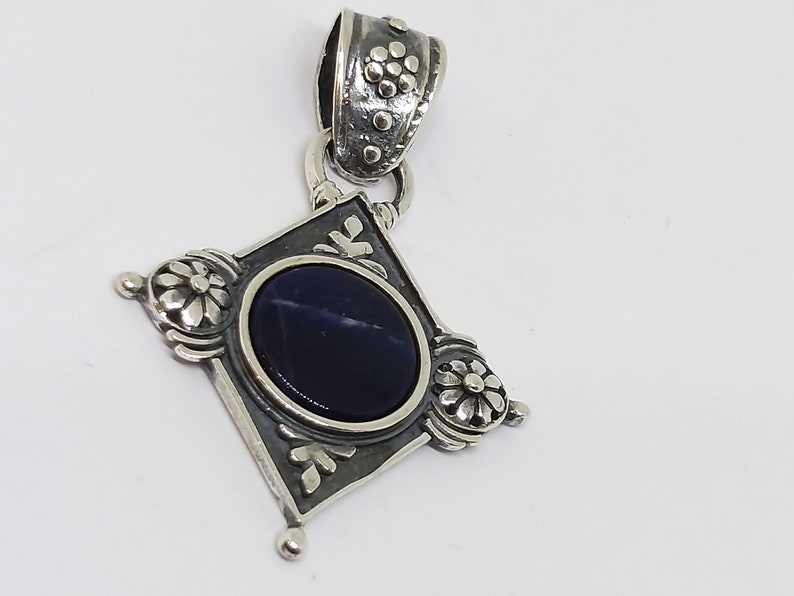 May include: A silver pendant with a dark blue gemstone set in a square frame with floral details. The pendant has a silver bail with a floral design.