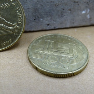 May include: Two gold-colored coins. The first coin features a person rowing a boat with the text "ATHENS 1997" and "100" visible. The second coin features a building with the text "100 &Delta;&Rho;&Alpha;&Chi;&Mu;&Epsilon;&Sigma;" visible.