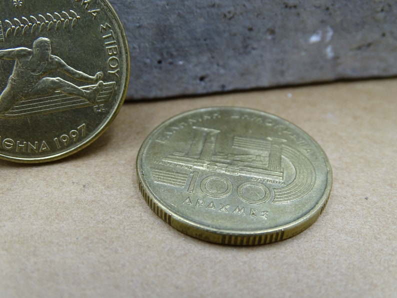 May include: Two gold-colored coins. The first coin features a person rowing a boat with the text "ATHENS 1997" and "100" visible. The second coin features a building with the text "100 &Delta;&Rho;&Alpha;&Chi;&Mu;&Epsilon;&Sigma;" visible.