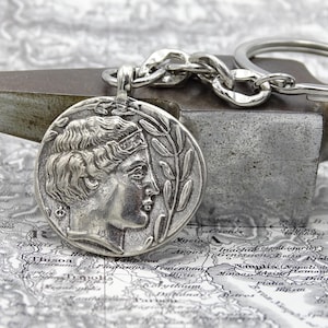 Puede incluir: Un llavero plateado con un colgante redondo con un diseño de perfil clásico. El colgante está unido a una cadena y un llavero. El fondo incluye un mapa y una superficie metálica.