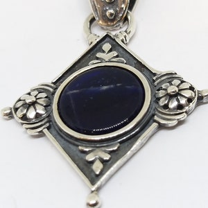 May include: A silver pendant with a dark blue gemstone set in a diamond-shaped frame. The frame is decorated with floral designs.