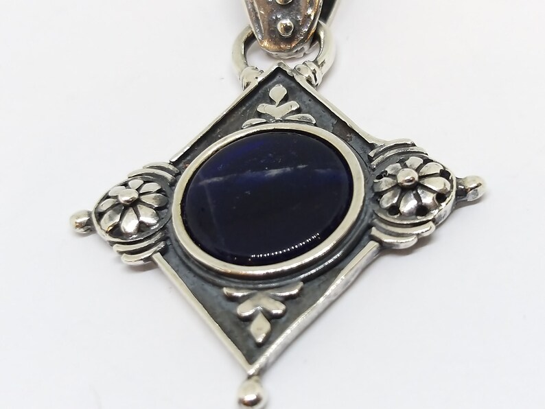 May include: A silver pendant with a dark blue gemstone set in a diamond-shaped frame. The frame is decorated with floral designs.