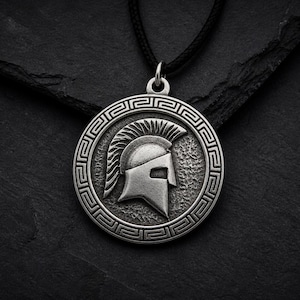 Può includere: Una collana con pendente color argento con un design a elmo spartano. Il pendente rotondo ha un bordo dettagliato ed è appeso a un cordino nero. Lo sfondo è una superficie scura e strutturata.