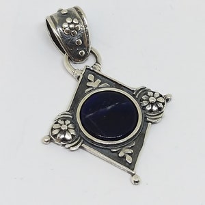 May include: A silver pendant with a dark blue gemstone set in a diamond-shaped frame. The frame is decorated with floral designs and the pendant has a silver bail.
