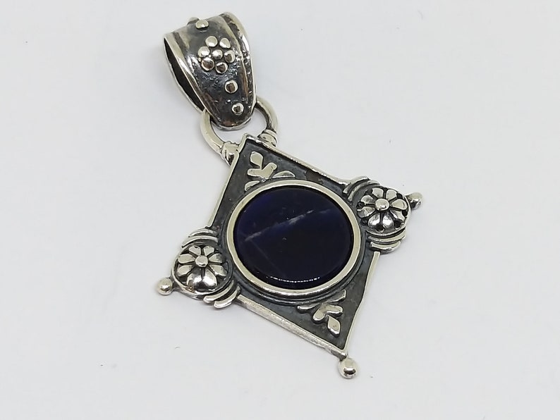 May include: A silver pendant with a dark blue gemstone set in a diamond-shaped frame. The frame is decorated with floral designs and the pendant has a silver bail.