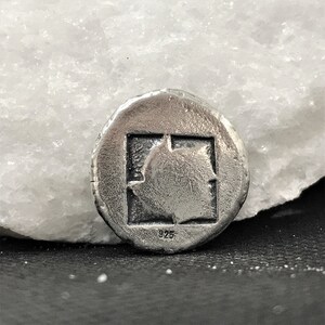 Puede incluir: Colgante redondo plateado con un dise&ntilde;o cuadrado central que contiene una forma estilizada de hoja o llama. El colgante est&aacute; marcado con "925", indicando la pureza del metal. Tiene un acabado texturizado y envejecido. Joyer&iacute;a.