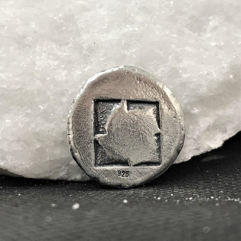 Puede incluir: Colgante redondo plateado con un dise&ntilde;o cuadrado central que contiene una forma estilizada de hoja o llama. El colgante est&aacute; marcado con "925", indicando la pureza del metal. Tiene un acabado texturizado y envejecido. Joyer&iacute;a.