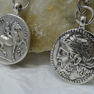 May include: Two silver-toned pendants with a chain. One pendant features a profile of a man with a laurel wreath, the other features a horse and rider.