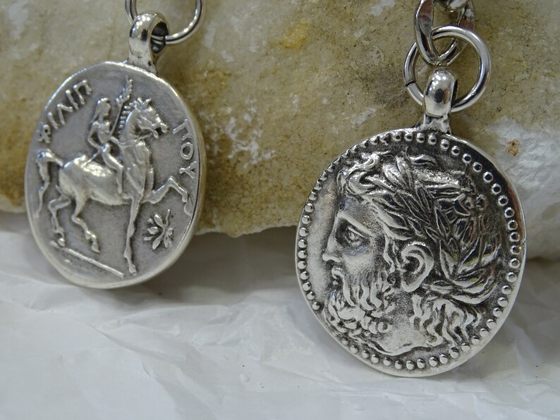 May include: Two silver-toned pendants with a chain. One pendant features a profile of a man with a laurel wreath, the other features a horse and rider.