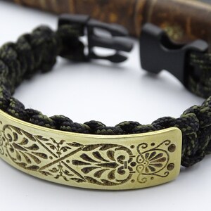 Puede incluir: Pulsera de paracord con cierre negro. La pulsera tiene un cord&oacute;n trenzado negro y verde oliva y una placa de lat&oacute;n con un dise&ntilde;o floral grabado. Un accesorio elegante, con una circunferencia de unos 18 cm.