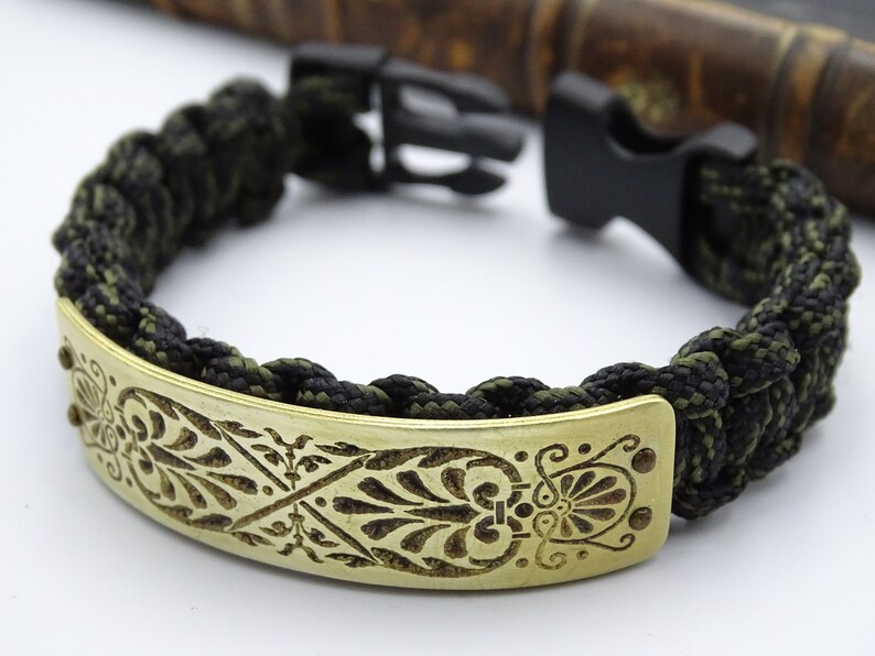 Puede incluir: Pulsera de paracord con cierre negro. La pulsera tiene un cord&oacute;n trenzado negro y verde oliva y una placa de lat&oacute;n con un dise&ntilde;o floral grabado. Un accesorio elegante, con una circunferencia de unos 18 cm.