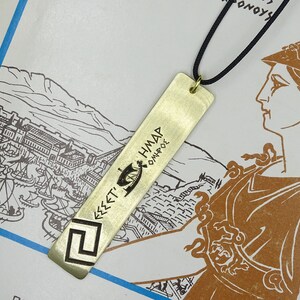 May include: A gold-coloured metal bookmark with a Greek inscription that reads "&Epsilon;&Sigma;&Sigma;&Epsilon;&Tau;&Iota; &Eta;&Mu;&Alpha;&Rho; &Omicron;&Mu;&Eta;&Rho;&Omicron;&Sigma;". The bookmark is hanging from a black cord and is on a white background with a drawing of a city.