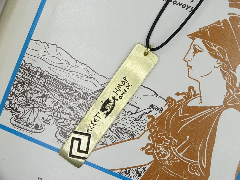 May include: A gold-coloured metal bookmark with a Greek inscription that reads "&Epsilon;&Sigma;&Sigma;&Epsilon;&Tau;&Iota; &Eta;&Mu;&Alpha;&Rho; &Omicron;&Mu;&Eta;&Rho;&Omicron;&Sigma;". The bookmark is hanging from a black cord and is on a white background with a drawing of a city.