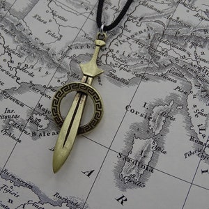 Może przedstawiać: Złoty wisiorek w kształcie miecza z okrągłym wzorem greckiego klucza wok&oacute;ł ostrza. Wisiorek jest zawieszony na czarnym sznurku.