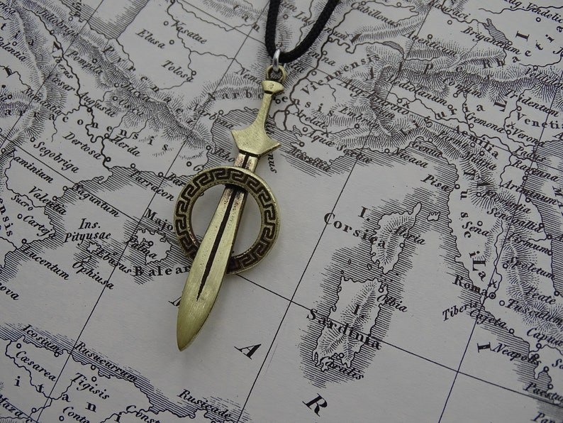 Może przedstawiać: Złoty wisiorek w kształcie miecza z okrągłym wzorem greckiego klucza wok&oacute;ł ostrza. Wisiorek jest zawieszony na czarnym sznurku.