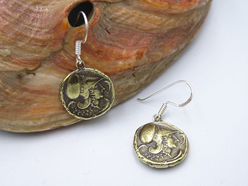May include: A pair of gold-toned earrings with a coin-shaped design featuring a profile of a woman's face. The earrings have silver hooks.