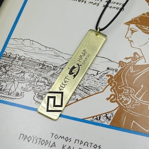 May include: A gold-coloured necklace with a rectangular pendant featuring a Greek key design and the inscription "&Epsilon;&Sigma;&Sigma;&Epsilon;&Tau;&Iota; &Eta;&Mu;&Alpha;&Sigma; &Omicron;&Iota;&Nu;&Omicron;&Pi;&Omicron;&Lambda;&Epsilon;" in ancient Greek. The pendant is hanging on a black cord.