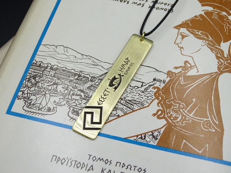 May include: A gold-coloured necklace with a rectangular pendant featuring a Greek key design and the inscription "&Epsilon;&Sigma;&Sigma;&Epsilon;&Tau;&Iota; &Eta;&Mu;&Alpha;&Sigma; &Omicron;&Iota;&Nu;&Omicron;&Pi;&Omicron;&Lambda;&Epsilon;" in ancient Greek. The pendant is hanging on a black cord.