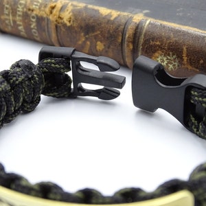 Puede incluir: Primer plano de una pulsera de paracord negra y verde oliva con un cierre de pl&aacute;stico negro. La pulsera est&aacute; hecha de un cord&oacute;n tejido apretado y presenta un acento met&aacute;lico de color dorado. Un libro vintage est&aacute; en el fondo.