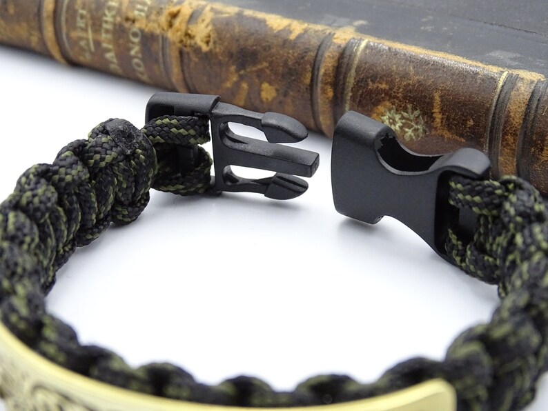 Puede incluir: Primer plano de una pulsera de paracord negra y verde oliva con un cierre de pl&aacute;stico negro. La pulsera est&aacute; hecha de un cord&oacute;n tejido apretado y presenta un acento met&aacute;lico de color dorado. Un libro vintage est&aacute; en el fondo.