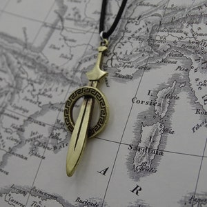 Może przedstawiać: Złoty wisiorek w kształcie miecza z okrągłym wzorem greckiego klucza wok&oacute;ł ostrza. Wisiorek wisi na czarnym sznurku na tle starej mapy.