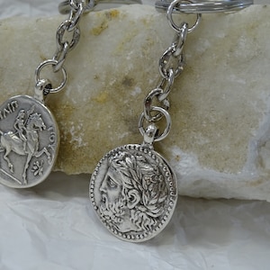May include: Two silver keychains with round coin-shaped charms. One charm features a rider on horseback, the other charm features a profile of a person with a laurel wreath.