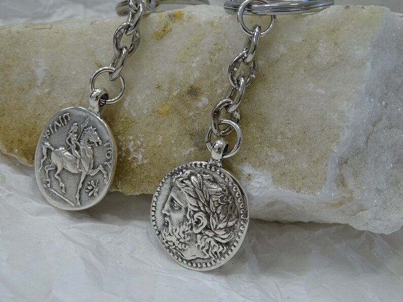 May include: Two silver keychains with round coin-shaped charms. One charm features a rider on horseback, the other charm features a profile of a person with a laurel wreath.