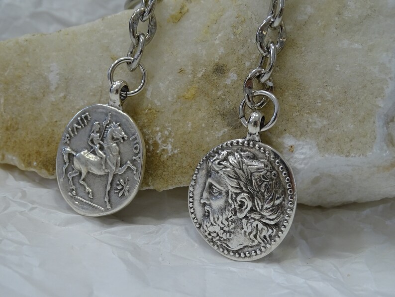 May include: Silver key chain with two round pendants. One pendant features a depiction of a rider on horseback with the text "&Pi;&Iota;&Nu;&Delta;&Alpha;&Rho;&Omicron;&Sigma;" and "&Iota;&Omicron;&Upsilon;&Sigma;". The other pendant features a profile of a man with a laurel wreath.