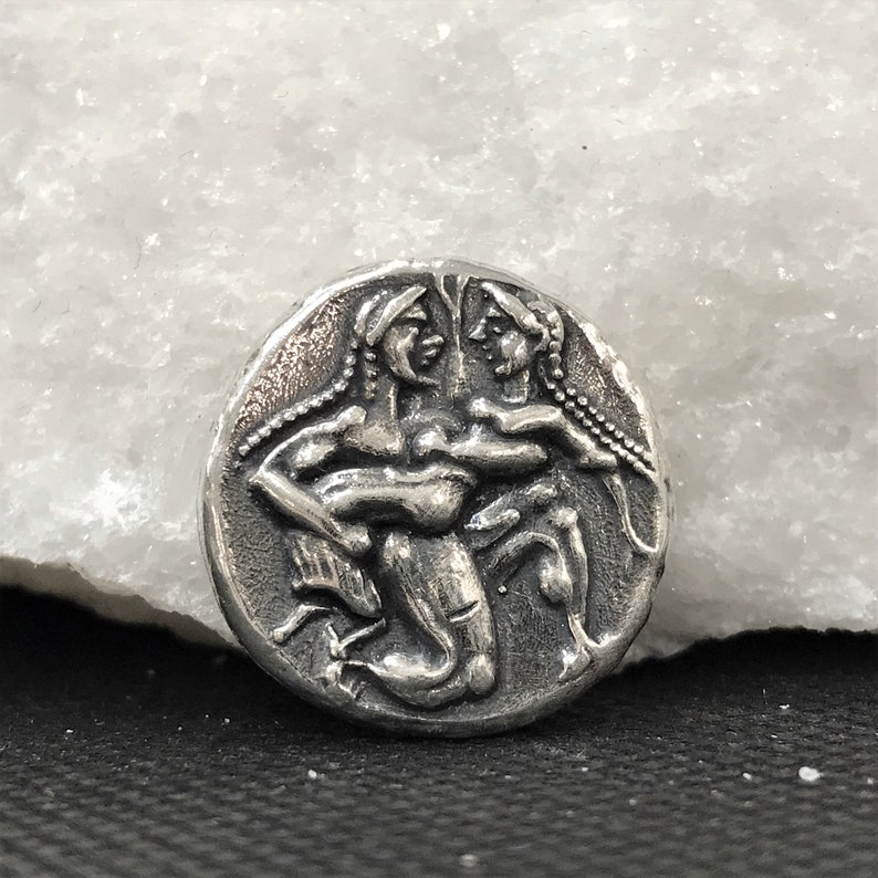 Puede incluir: Una moneda redonda, de color plateado, con un relieve detallado de dos figuras abraz&aacute;ndose. Las figuras tienen pelo largo y est&aacute;n representadas en un estilo cl&aacute;sico. La moneda est&aacute; sobre un fondo blanco y negro.