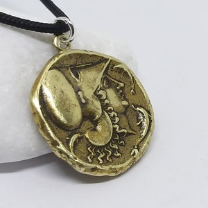 K&ouml;nnte beinhalten: Eine goldfarbene Halskette mit einem detaillierten Profil eines Frauenkopfes mit einer Krone. Der Anh&auml;nger ist rund und hat eine strukturierte Oberfl&auml;che. Die Halskette ist an einer schwarzen Schnur.