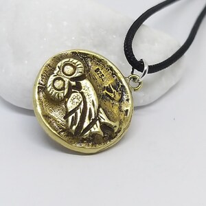 May include: A gold-colored pendant necklace featuring an owl design. The pendant is round and has a textured surface. The necklace is on a black cord.