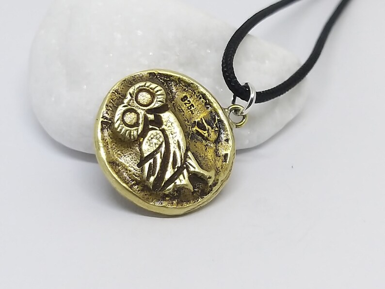 May include: A gold-colored pendant necklace featuring an owl design. The pendant is round and has a textured surface. The necklace is on a black cord.