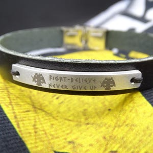 May include: Black leather bracelet with a silver plate engraved with the words "FIGHT-BELIEVE NEVER GIVE UP" and two stylized eagle emblems. The bracelet has a silver clasp and a green interior.