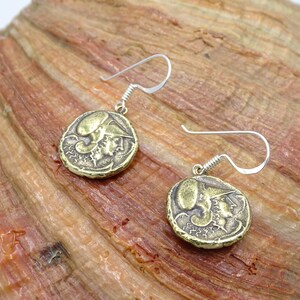 May include: A pair of gold-toned earrings featuring a round coin design with a raised image of a person's head. The earrings have silver-toned hooks.