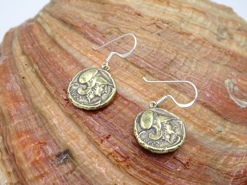 May include: A pair of gold-toned earrings featuring a round coin design with a raised image of a person's head. The earrings have silver-toned hooks.